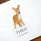 A close-up of a personalized white textured card featuring a watercolor illustration of a young brown deer and the printed name 'THEO' with the date '03.09.