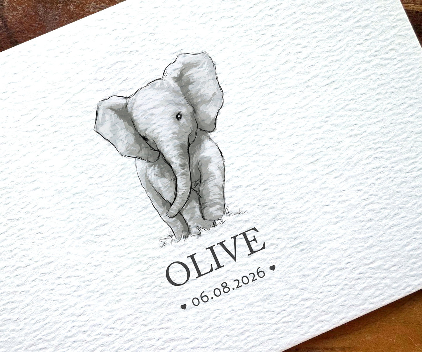 A close-up of a white textured paper card featuring a black-and-white sketch of a baby elephant above the name 'OLIVE' and the date 06.08.2026.